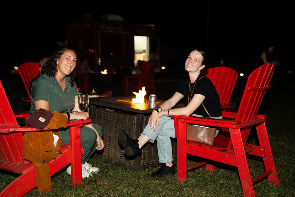 Two women sitting in red Adirondack chairs near a fire pit smile for a photo