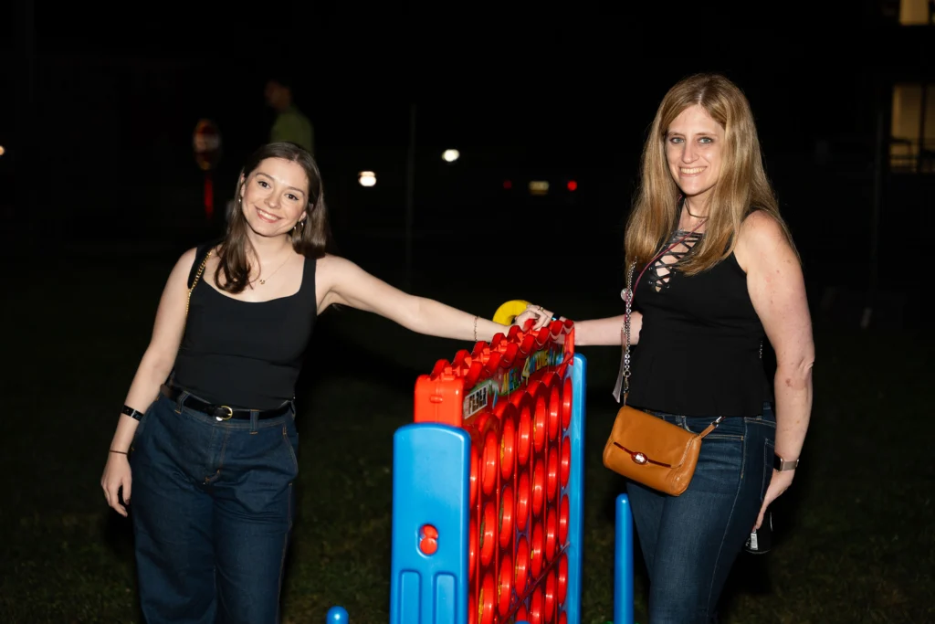 Two women in black shirts smile next to a game of giant Connect 4