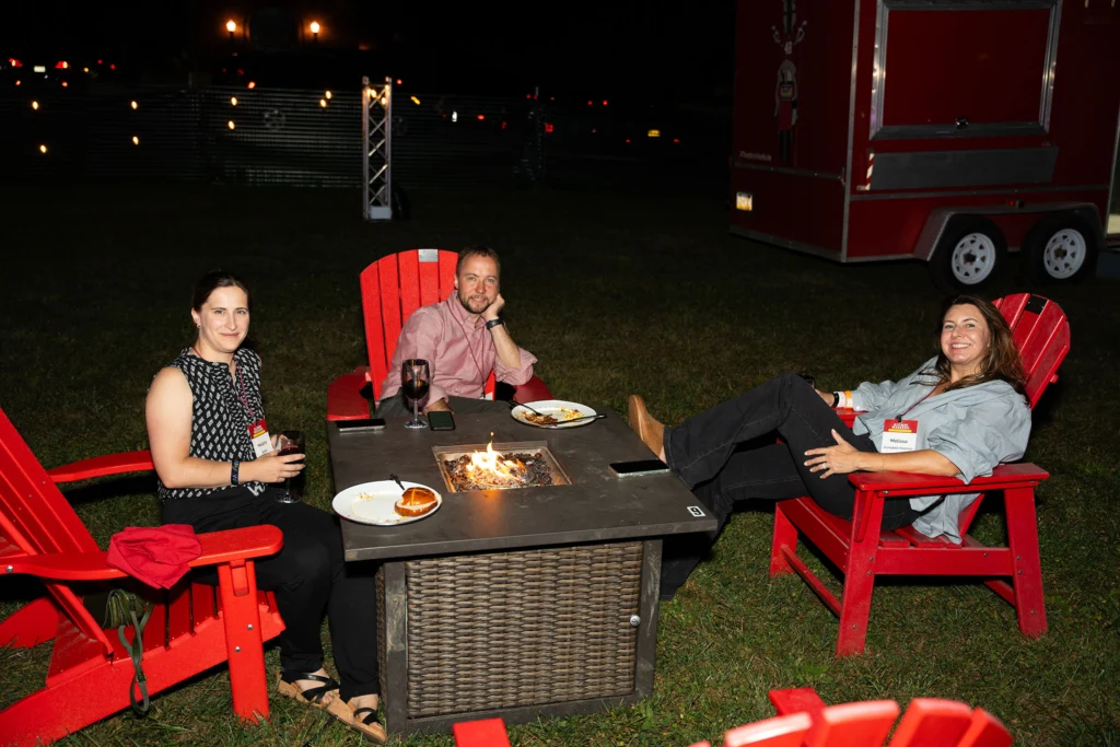 Three people sit in red Adirondack chairs around a fire pit