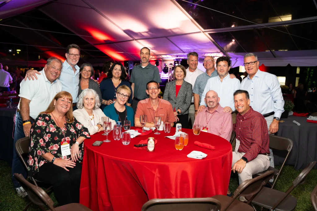 A large group of people smile for a photo around a table with a red tablecloth under a tent