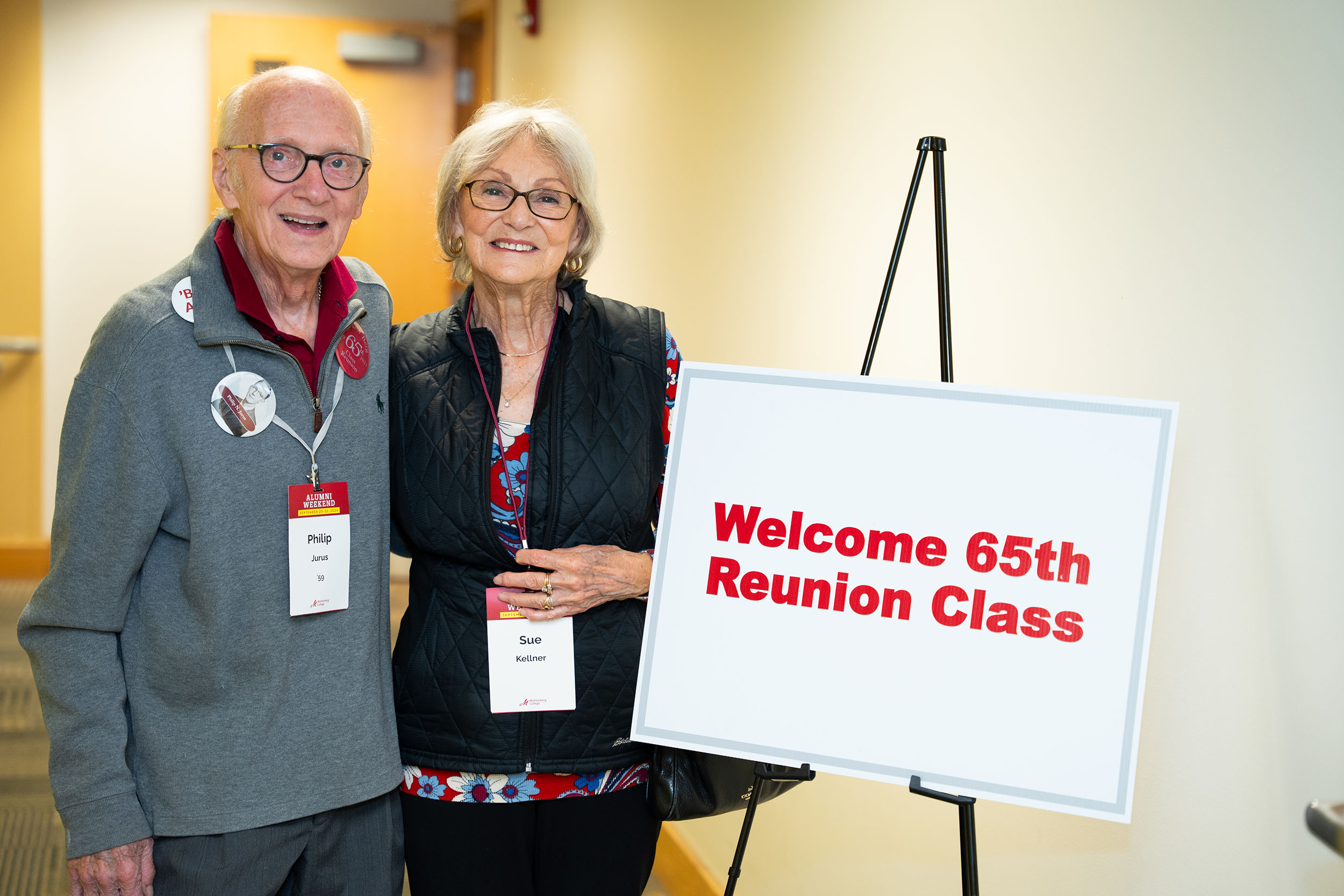 Two people stand next to a sign that says Welcome 65th Reunion Class