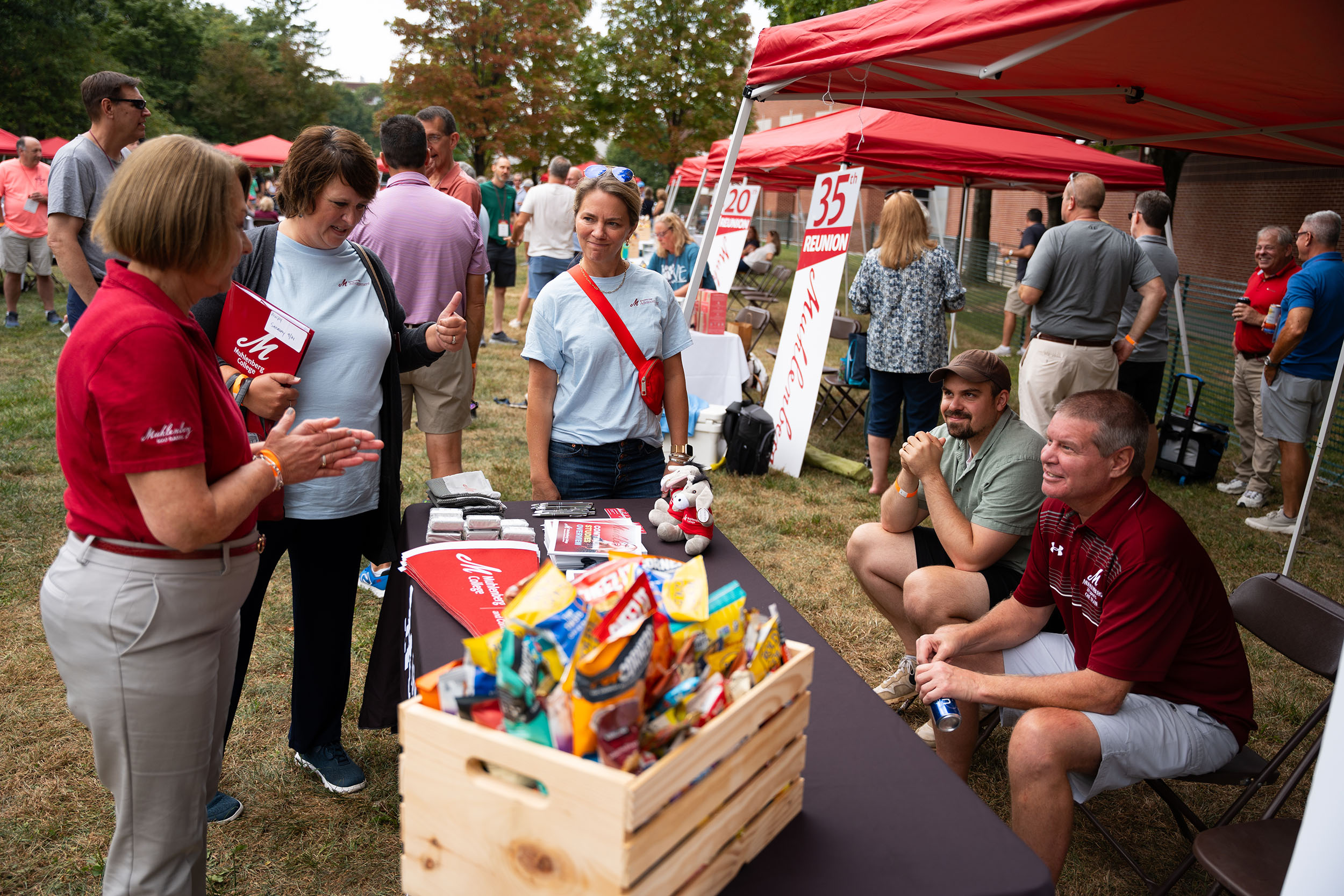 People stand around a table outside and talk with tents in the background that have signs reading 35th Reunion and 20th Reunion