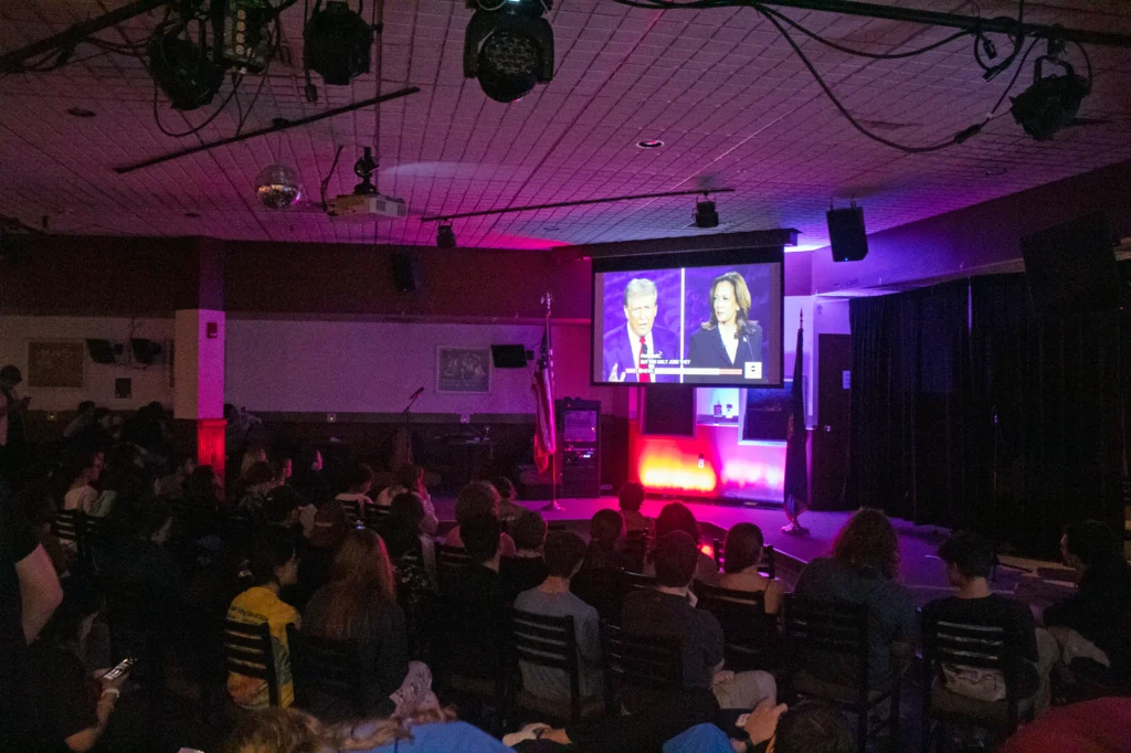 A dark room filled with people looking at a projector screen with the presidential debate on it.