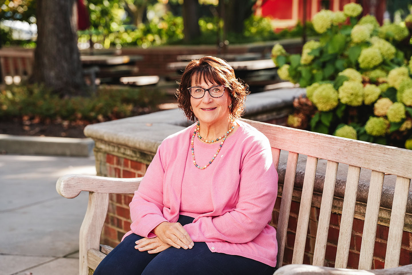 A woman in a pink sweater and glasses poses for a photo on a bench