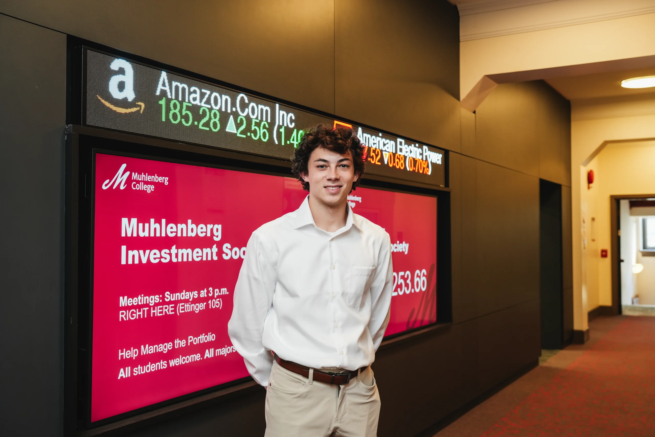 A college student stands in front of a stock ticker and video board that says Muhlenberg Investment Society