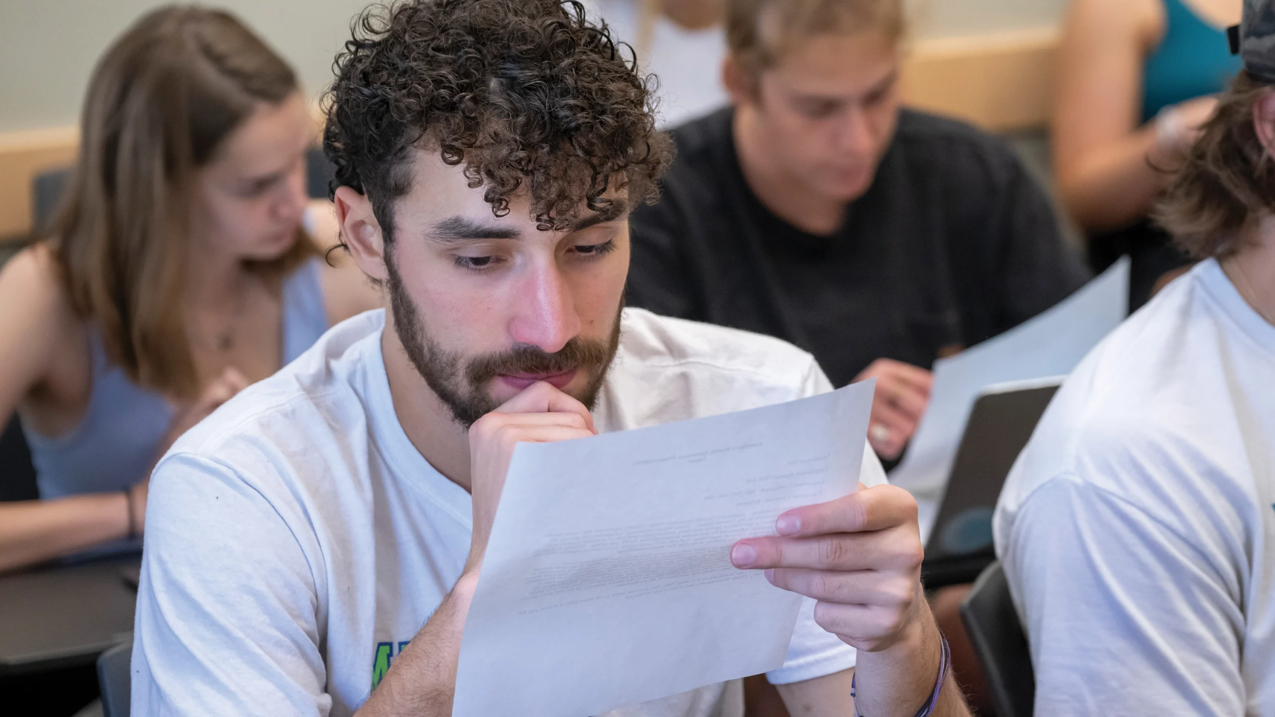 Photo of a male student looking at a paper with other students in the background.