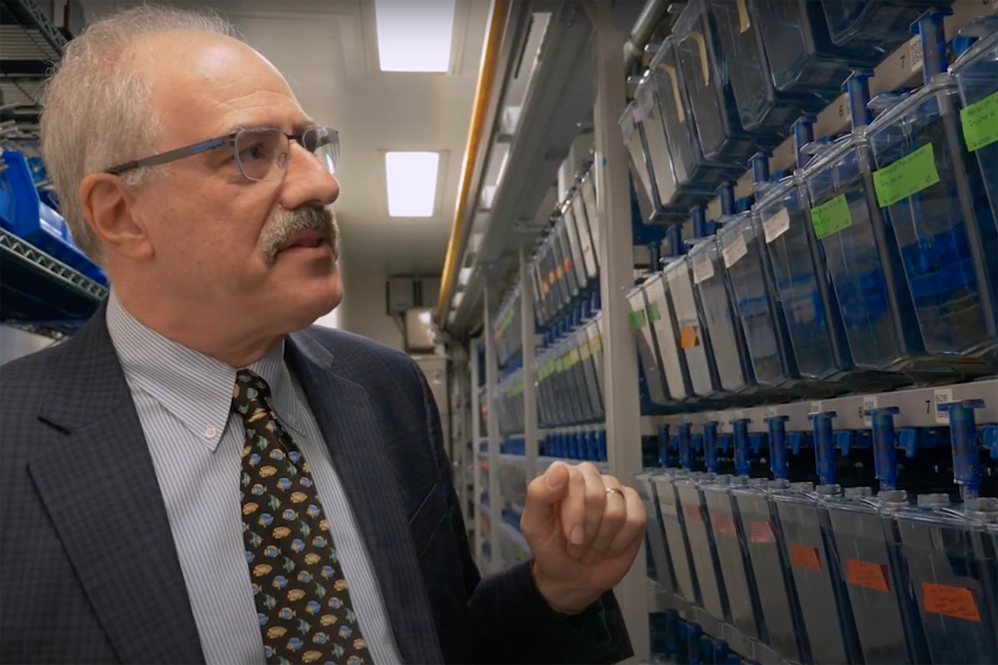 A scientist in a suit and tie stands in a room full of fish tanks