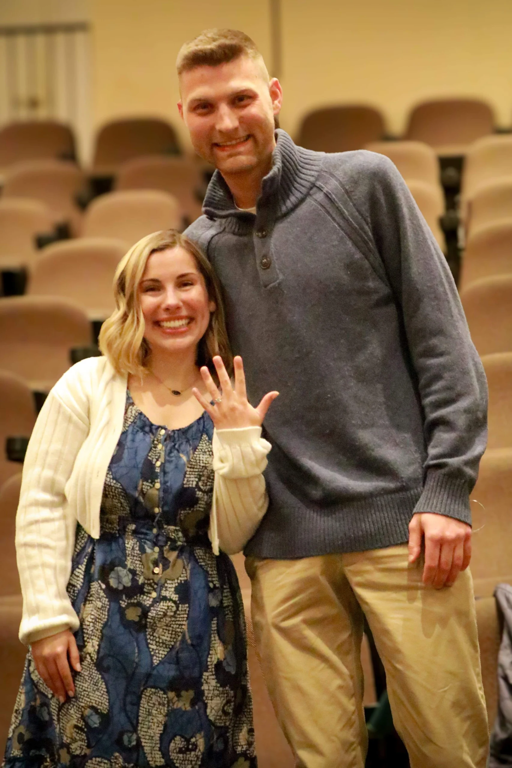 Two young people pose for a photo, the woman holds up her hand to show a ring