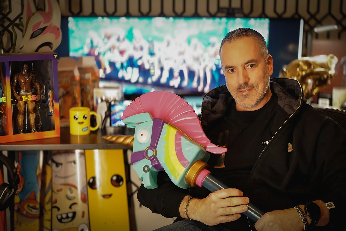 A man holds a unicorn head on a stick and sits in front of Fortnite memorabilia