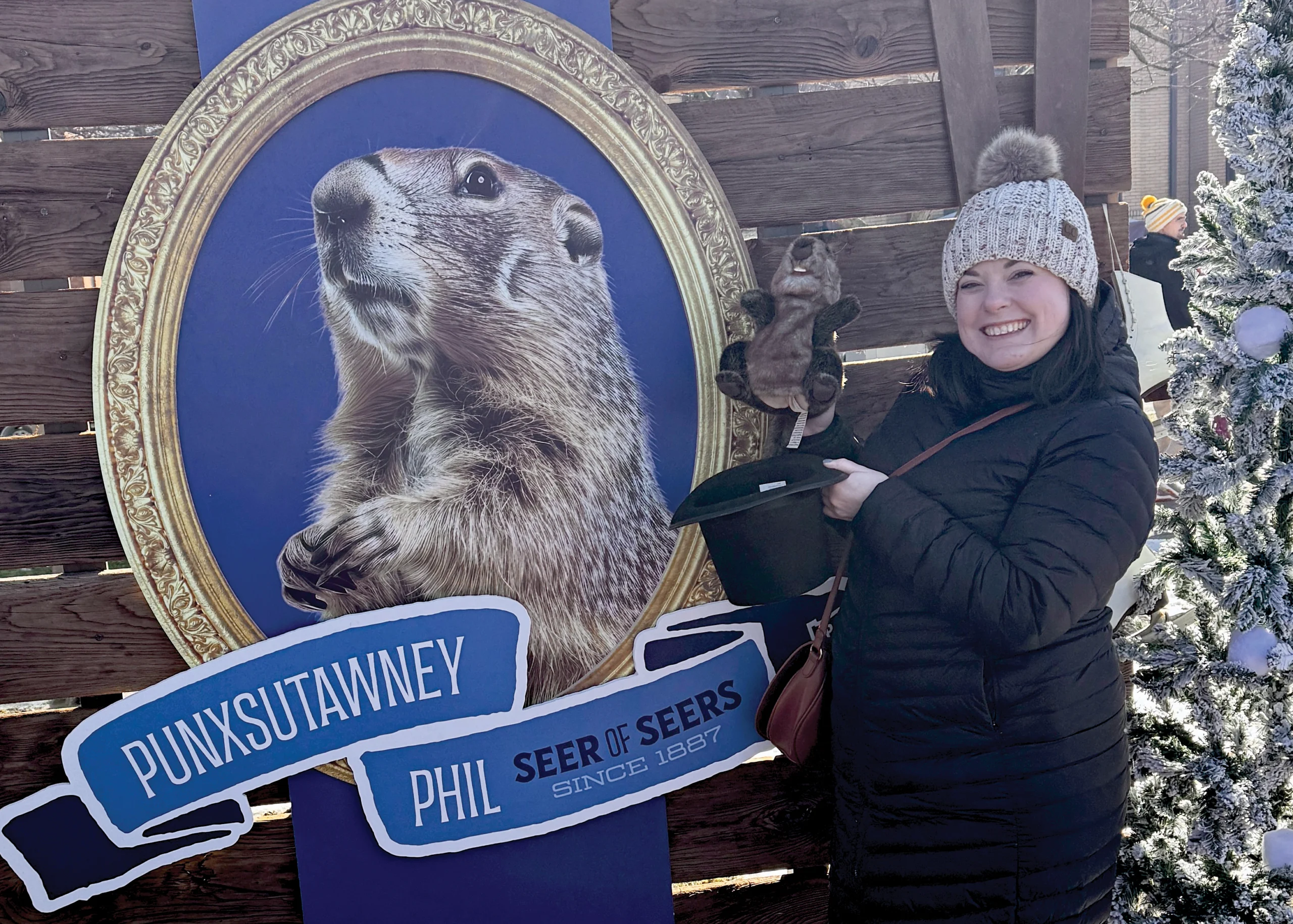 A photo of Kaitie Burger standing to the right of a Punxsutawney Phil poster smiling while holding a black top hat and groundhog stuffed animal