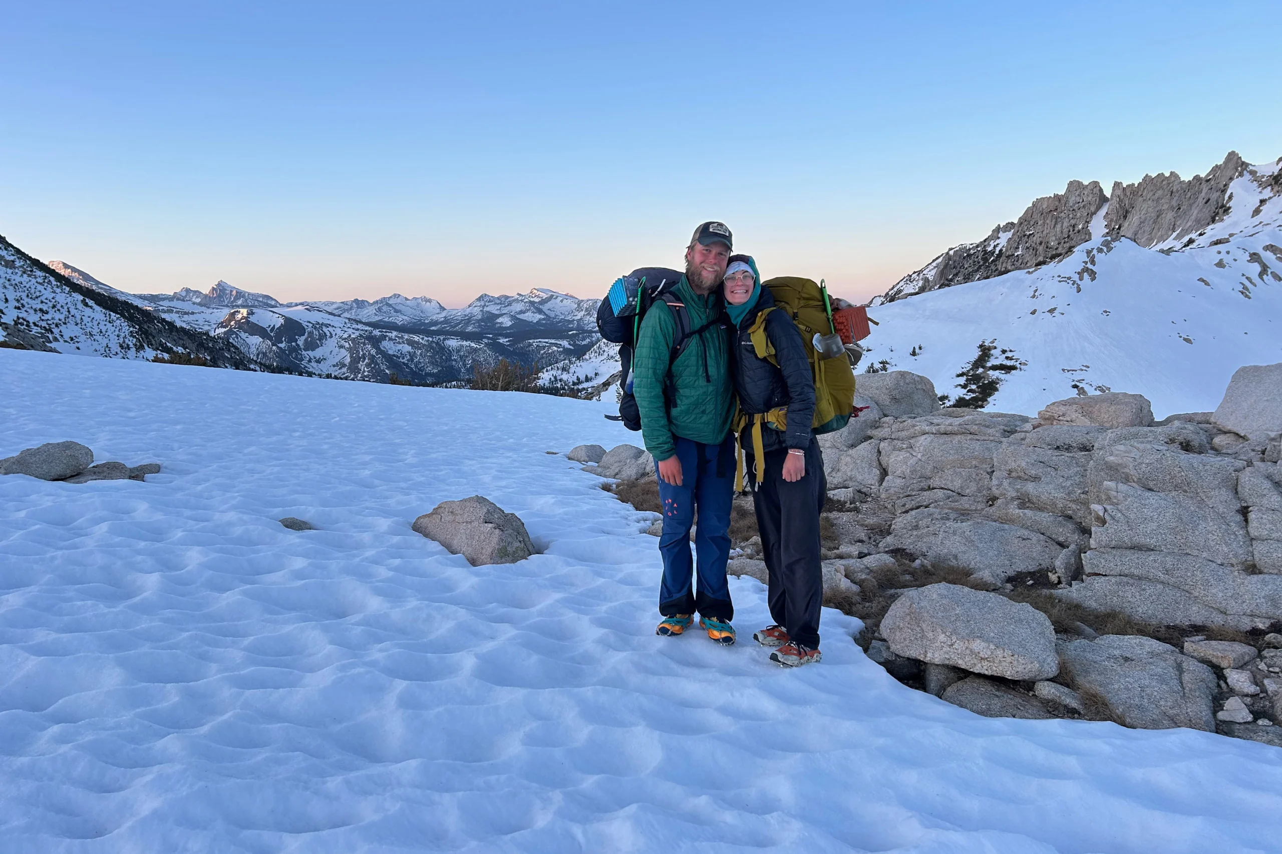 Two hikers wearing large packs smile while standing in snow in the mountains