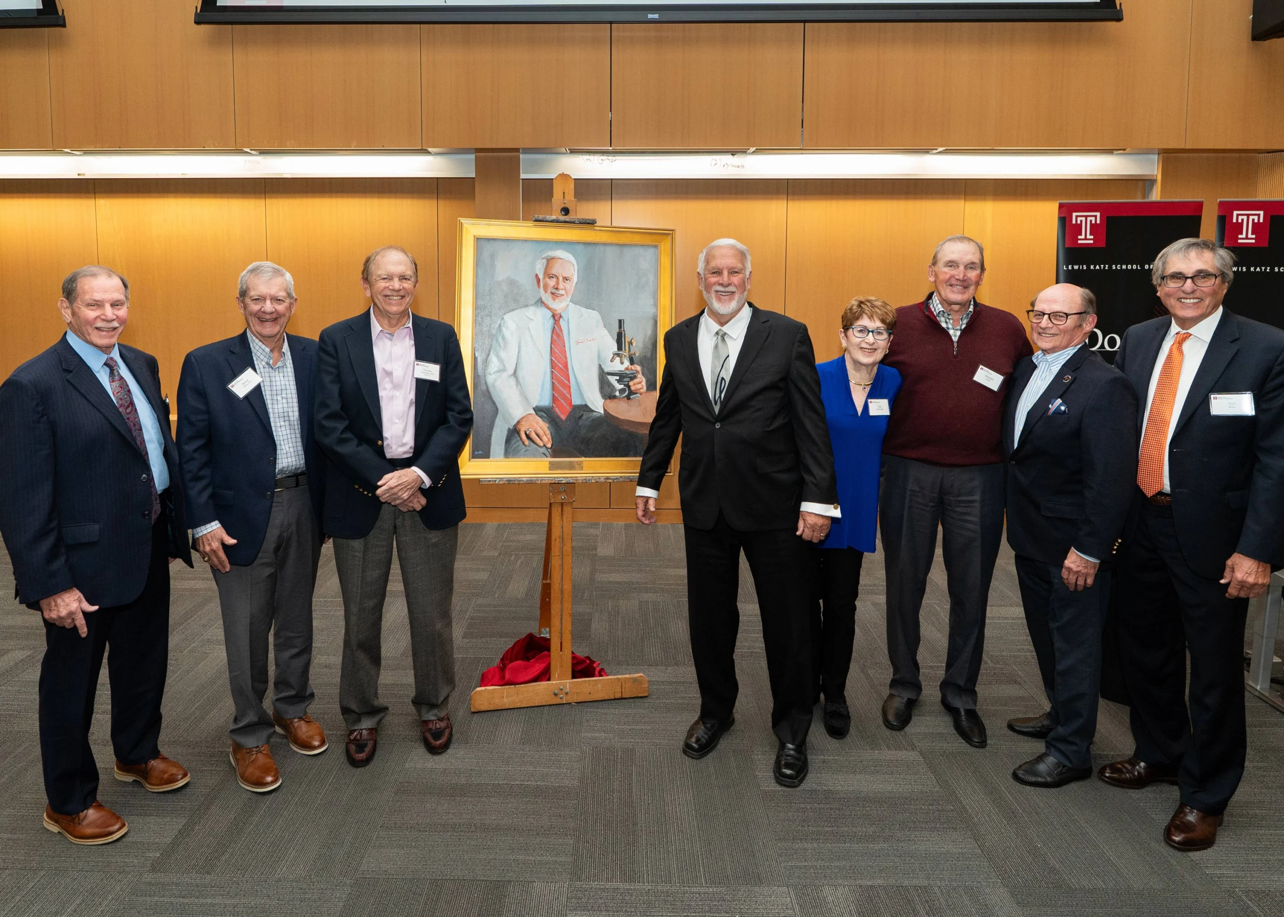 A group of adults in suits poses with a painted portrait of the man in the center