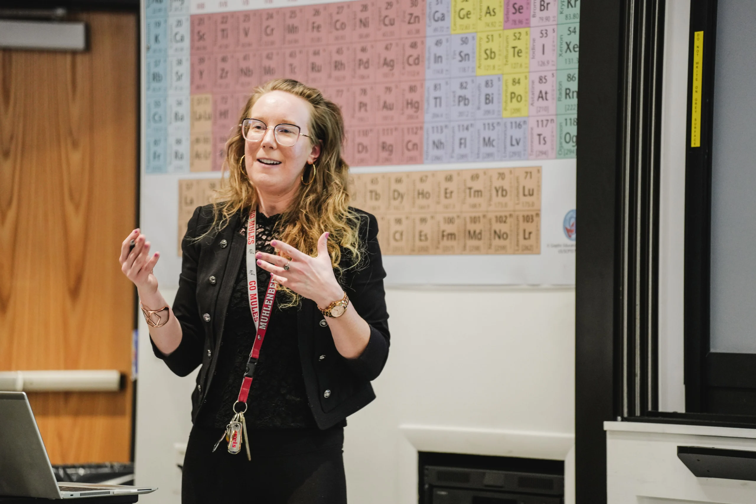 A college professor with curly hair and glasses lectures in front of a periodic table